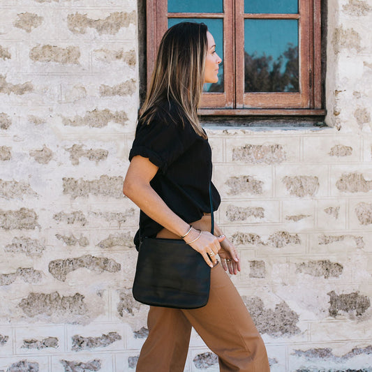Woman wearing a black top and brown pants standing against a textured wall with a window with a black leather crossbody bag.