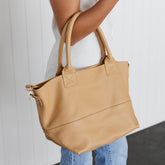 A woman holding a beige leather tote bag over their forearm against a white background.