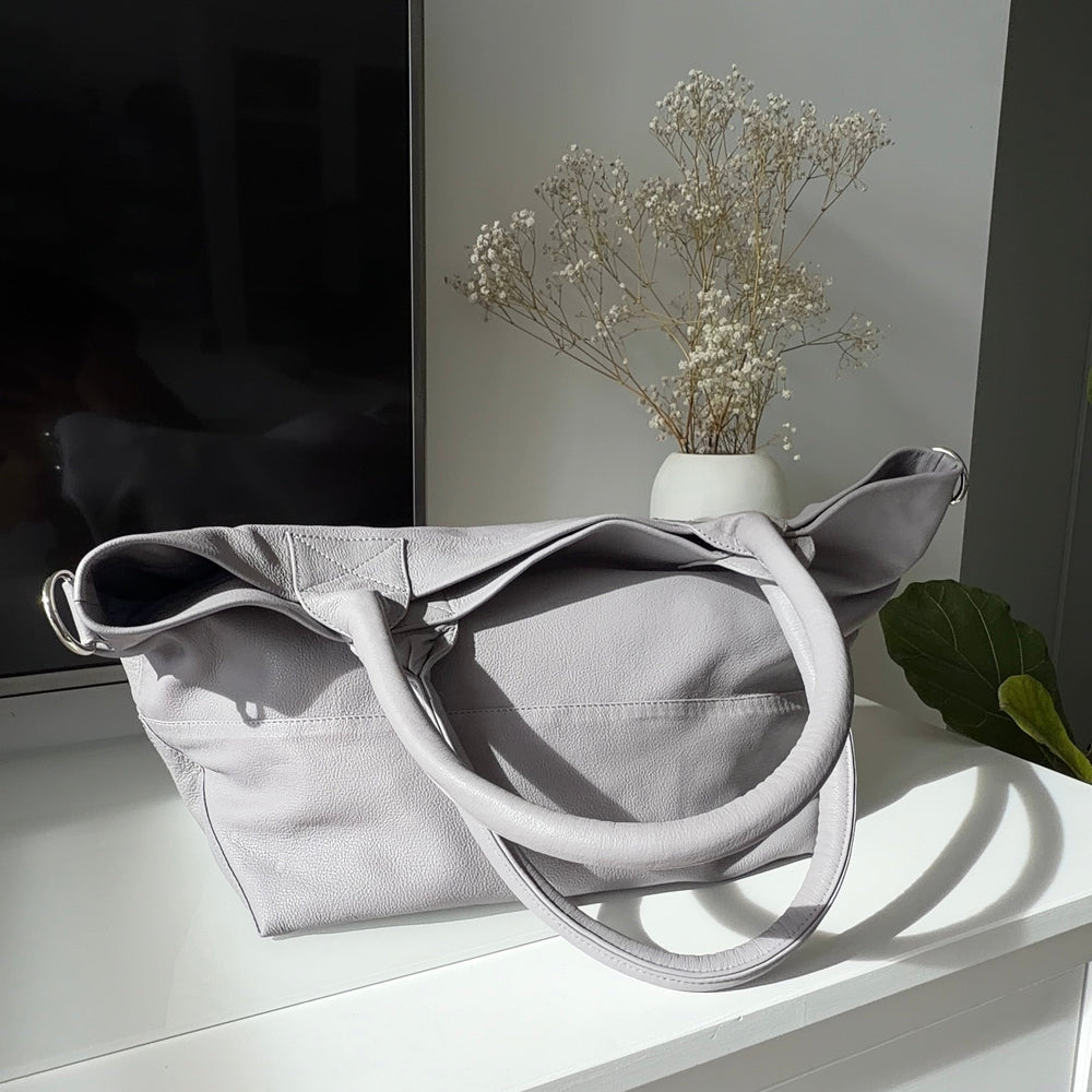 Grey leather tote bag on a white surface with a vase of dried flowers in the background
