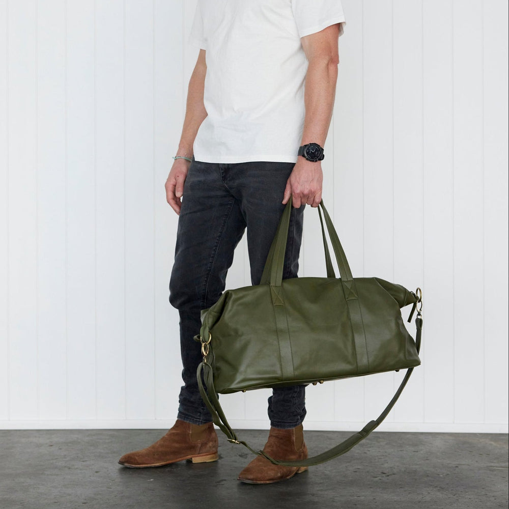 A man wearing a white t-shirt, dark jeans, and brown boots stands in a studio holding a large army
green leather duffel bag.