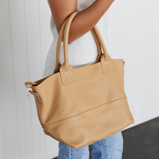 A woman holding a beige leather tote bag over their forearm against a white background.