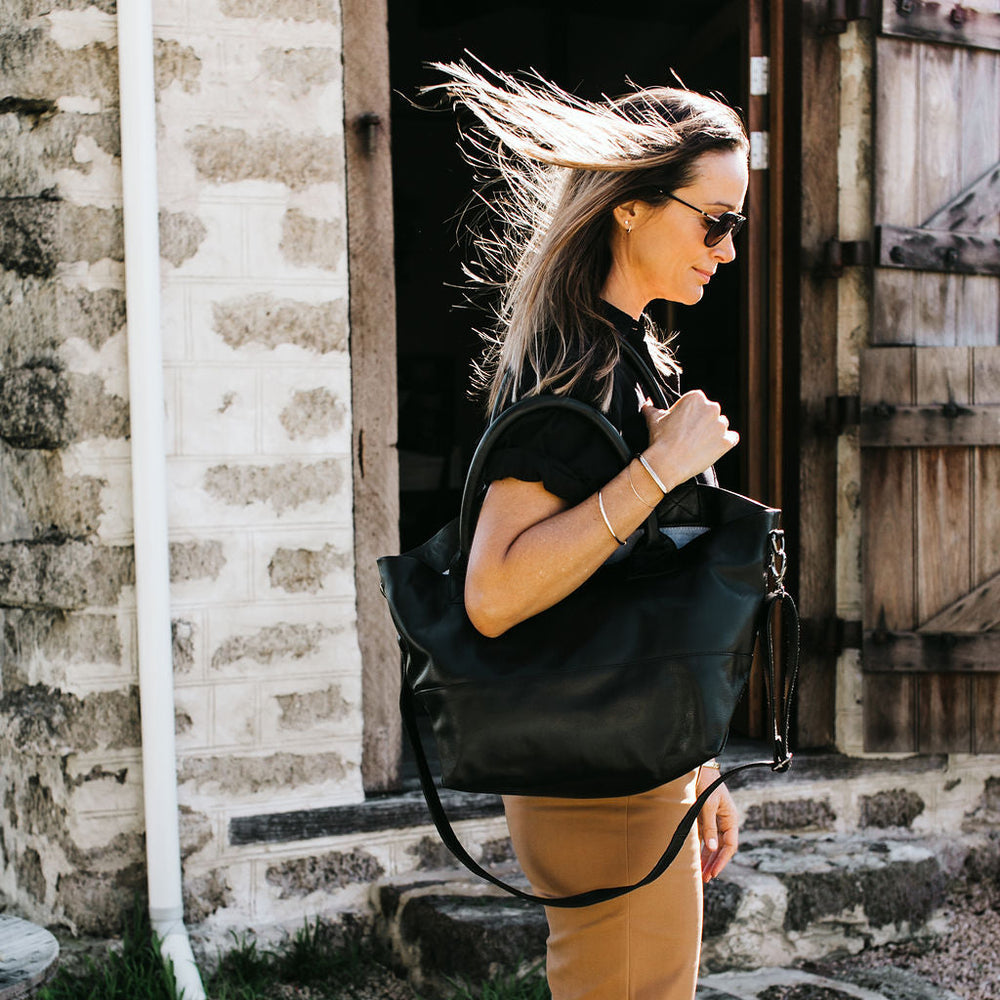 Woman holding a black leather 
tote bag outdoors with a rustic background