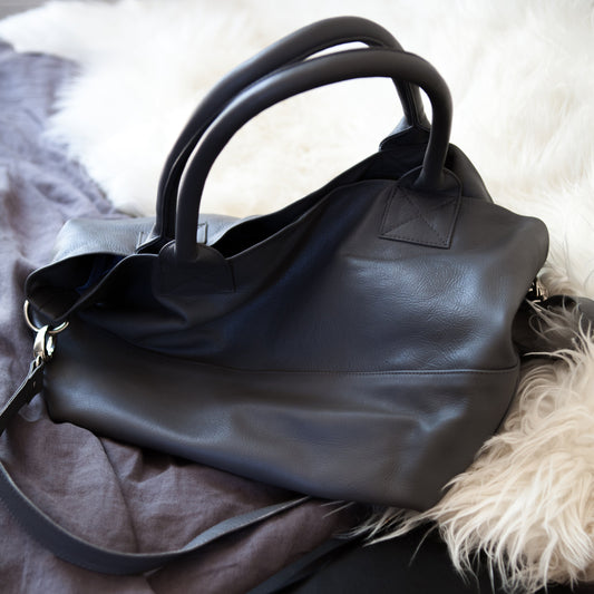 Dark grey leather handbag on a textured surface with a notebook and glasses.