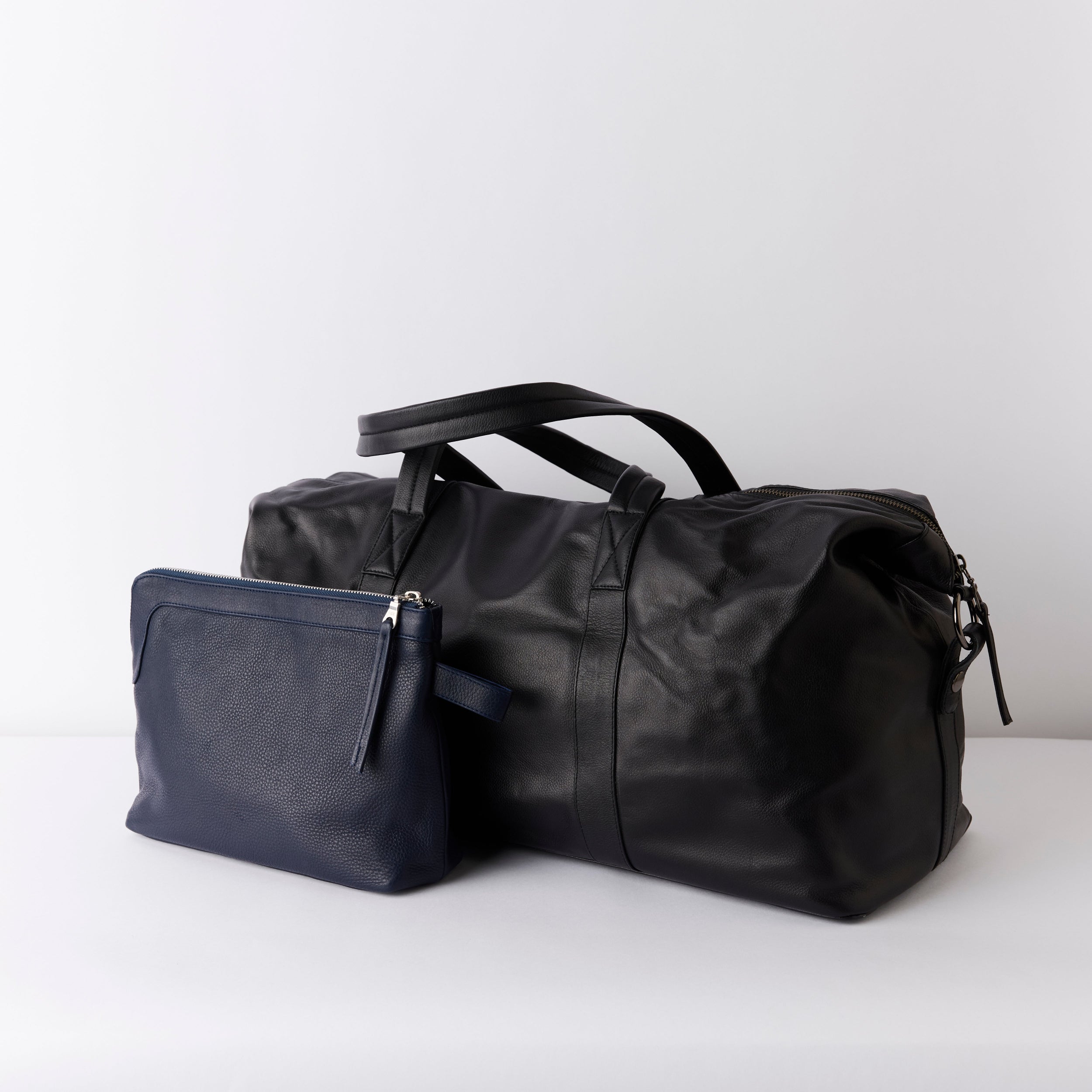 Black leather weekender / duffel bag and Navy toiletry bag on a white background