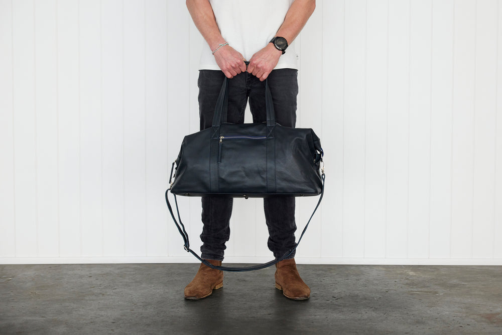 A person standing against a white wall holds a large french navy leather weekender / duffel bag with a long shoulder strap. The person is wearing a white t-shirt, dark jeans, and brown suede boots.
