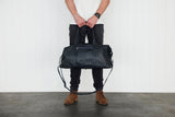 A person standing against a white wall holds a large french navy leather weekender / duffel bag with a long shoulder strap. The person is wearing a white t-shirt, dark jeans, and brown suede boots.