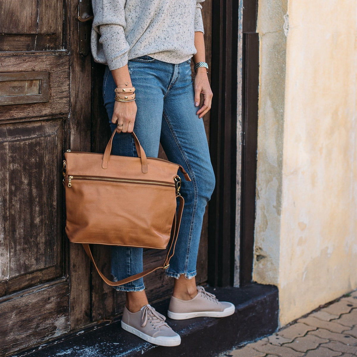 A person standing by a wooden door, wearing a grey sweater and blue jeans, carrying a Rome Caramel brown leather tote bag with a long strap.